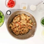 Large mixing bowl of shredded chicken with diced vegetables, surrounded by bowls of lettuce, chopped tomato, cilantro, seasoning, lime juice, and a whole avocado on a white surface&mdash;perfect for assembling bold and flavorful Firecracker Chicken Wraps.