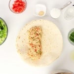 Firecracker Chicken Wraps featuring a tortilla with chicken filling in the center, surrounded by bowls of lettuce, tomatoes, herbs, spices, sugar, oil, avocado, and lemon juice on a white surface.