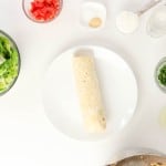 A burrito sits on a white plate surrounded by bowls of lettuce, diced tomato, cilantro, avocado, seasonings, and other ingredients on a white background&mdash;perfect fixings to create your own vibrant Firecracker Chicken Wraps.