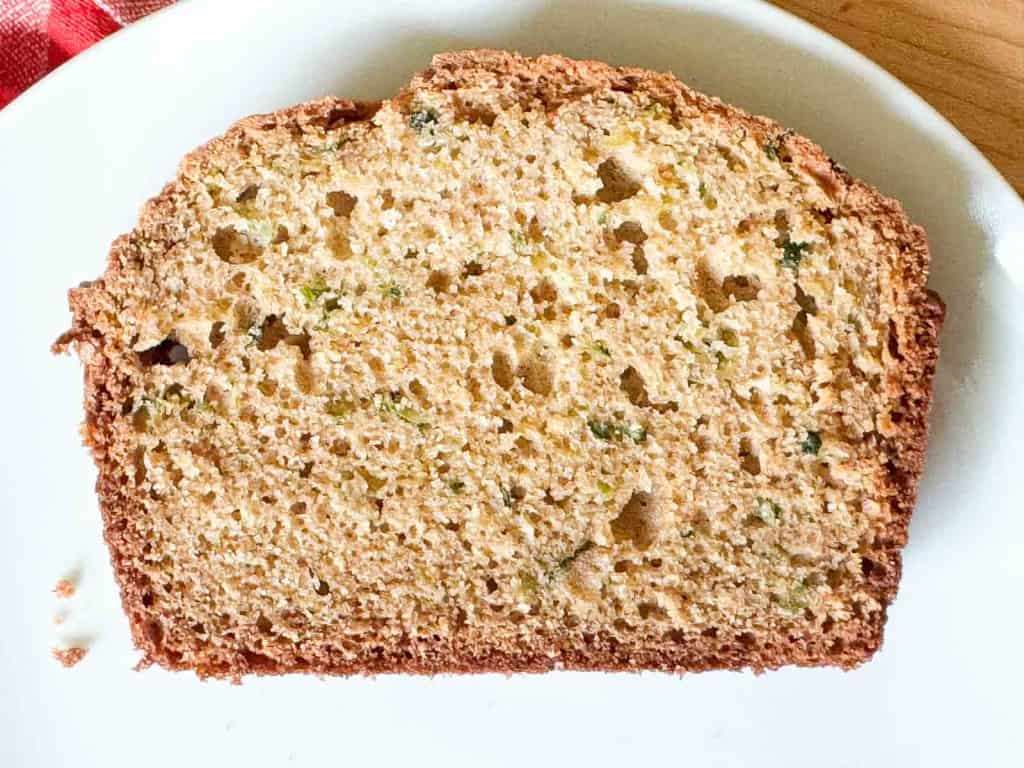 A single slice of zucchini bread rests on a white plate, displaying a golden-brown crust and a moist, speckled zucchini bread interior.
