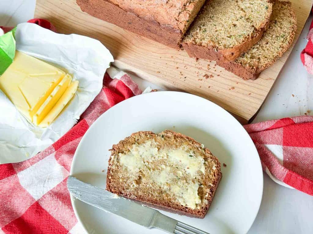 A plate with a buttered slice of zucchini bread, a knife, and a red checkered cloth. Sliced loaf and butter are on a wooden board in the background.