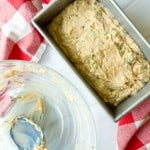 A loaf pan filled with raw zucchini bread batter sits next to a glass bowl with remnants of zucchini bread batter and a spatula, on a white surface with a red checkered cloth.