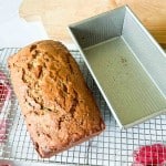 A loaf of banana bread sits on a cooling rack next to an empty metal loaf pan, with a wooden cutting board in the background&mdash;perfectly capturing the comforting scene often found when baking classics like zucchini bread.