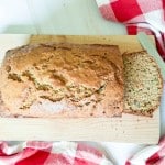 A loaf of homemade zucchini bread with one slice cut, placed on a wooden board next to a serrated knife and a red-and-white checkered cloth.