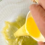 A hand pours melted butter from a small white cup into a bowl containing a yellow-green liquid.
