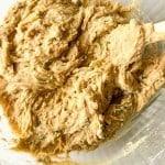 Close-up of a thick, beige batter with green specks in a clear glass mixing bowl, being stirred with a white spatula.