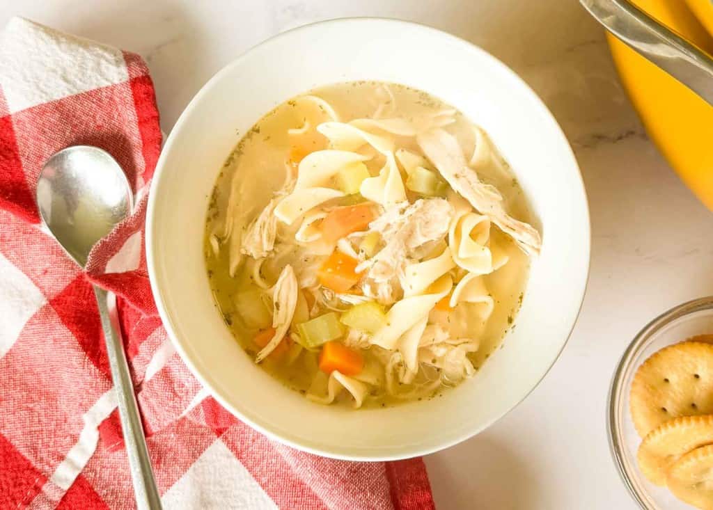A bowl of easy chicken noodle soup with carrots and celery sits on a table next to a spoon, a red and white napkin, and a small dish of round crackers.