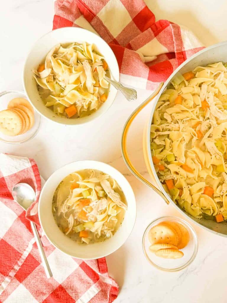 Overhead view of two bowls and a pot of easy chicken noodle soup with crackers on the side, set on a table with red and white checkered napkins.