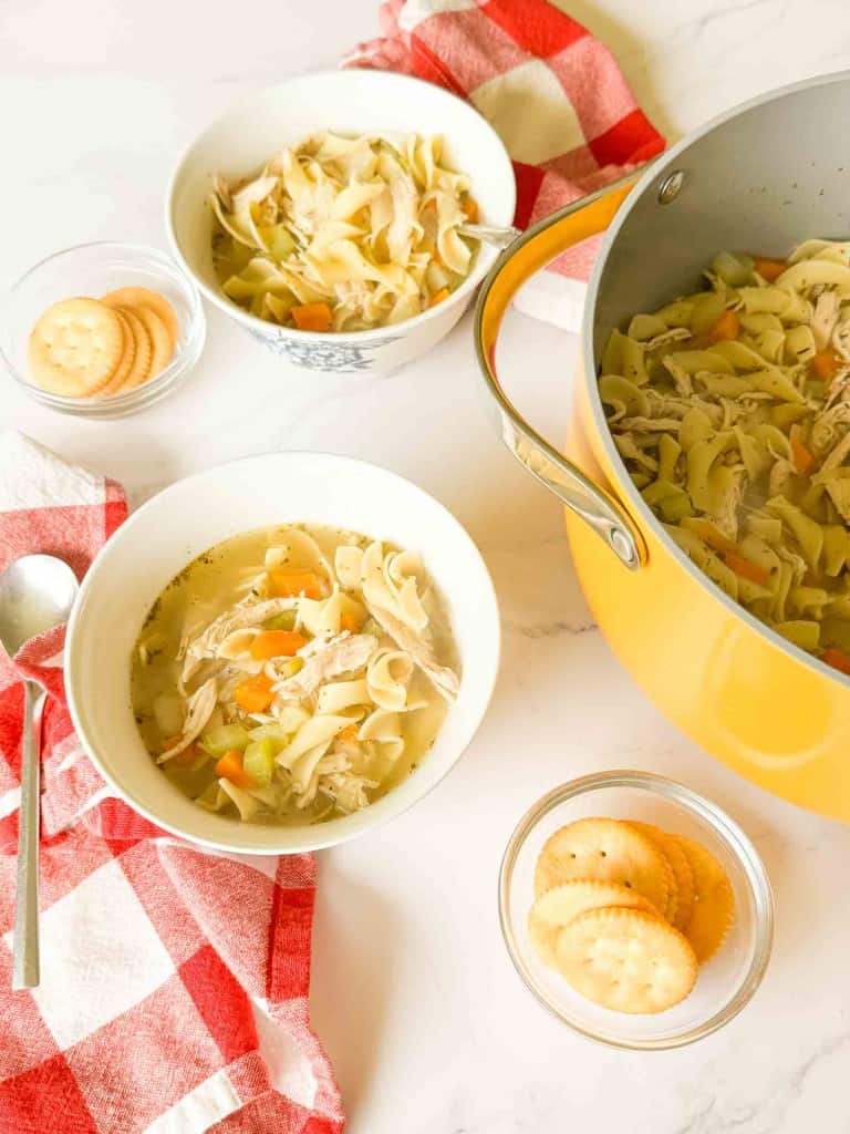 A pot of easy chicken noodle soup with carrots and noodles is served in two bowls, each with a spoon, next to bowls of round crackers on a red and white checkered towel.