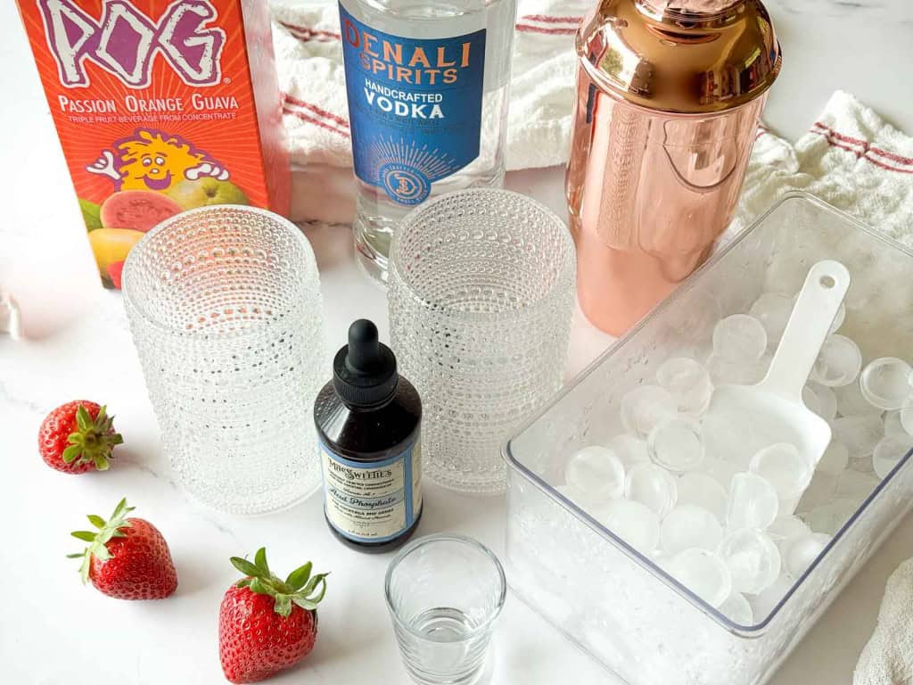 Box of juice, bottle of vodka, cocktail shaker, three glass tumblers, a container of ice with a scoop, strawberries, bitters, and a shot glass arranged on a countertop.