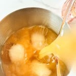 A pitcher pours orange liquid over ice cubes in a metal bowl on a white countertop.