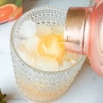 A drink is being poured from a shaker into a glass filled with ice cubes on a white surface. A box with a fruit image and some green leaves are visible nearby.