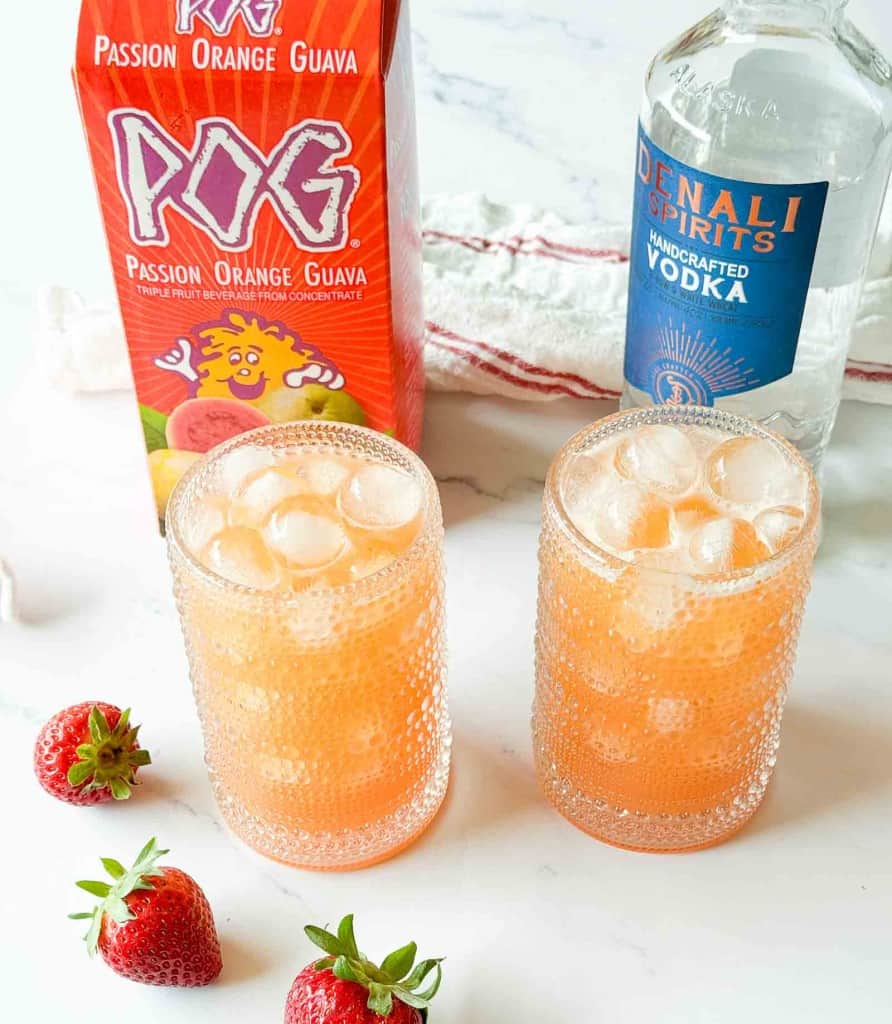 Two glasses filled with ice and an orange drink are placed beside a carton of POG juice, a vodka bottle, and several strawberries on a white surface.