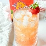 A glass of iced passion orange guava juice garnished with a strawberry, with a juice carton and a towel in the background.
