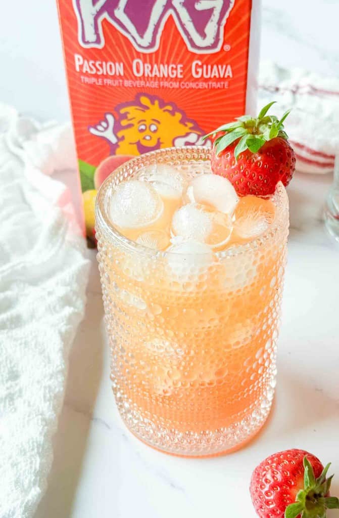 A glass of iced passion orange guava juice garnished with a strawberry, with a juice carton and a towel in the background.