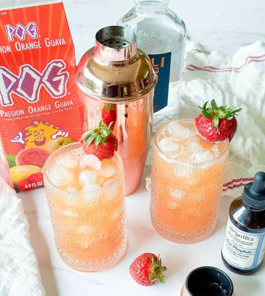 Two iced cocktails garnished with strawberries beside a juice box, a cocktail shaker, a liquor bottle, a bitters bottle, and a towel on a marble surface.