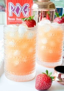 Two textured glasses filled with an orange-pink iced drink topped with strawberries, with a cocktail shaker and POG juice carton in the background.
