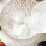 Round ice cubes being poured into a textured glass bowl, with part of a strawberry and a juice carton visible nearby.
