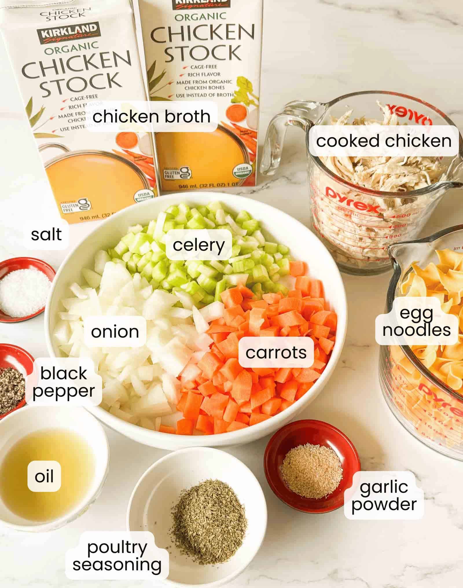 Ingredients for easy chicken noodle soup are arranged on a counter, including chicken broth, cooked chicken, egg noodles, diced celery, carrots, onion, and various seasonings in small bowls.