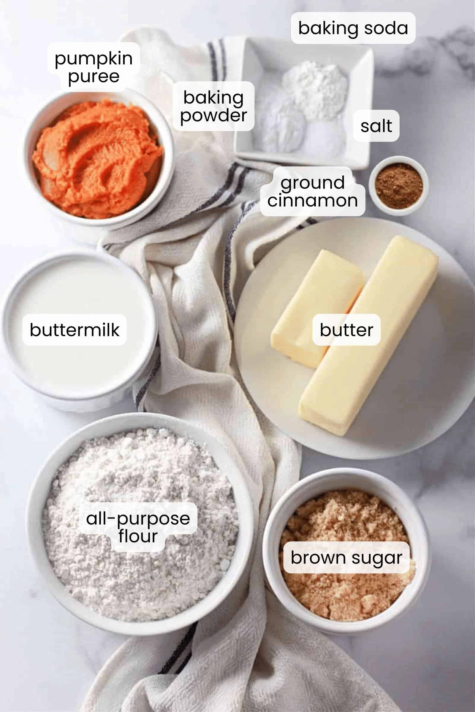 Assorted baking ingredients in bowls on a white surface, including flour, brown sugar, buttermilk, butter, pumpkin puree, baking powder, baking soda, salt, and ground cinnamon—perfect for making soft and flavorful pumpkin buttermilk biscuits.