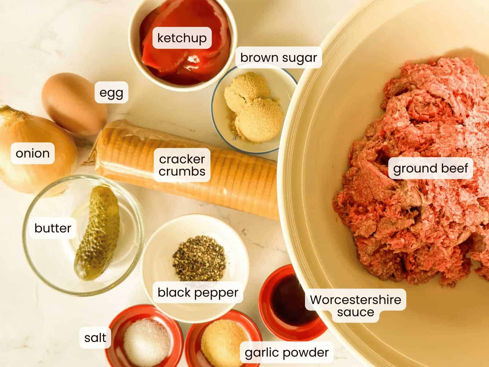 Top-down view of labeled slow cooker meatloaf ingredients: ground beef, cracker crumbs, onion, egg, butter, pickle, ketchup, brown sugar, Worcestershire sauce, garlic powder, salt, and black pepper.