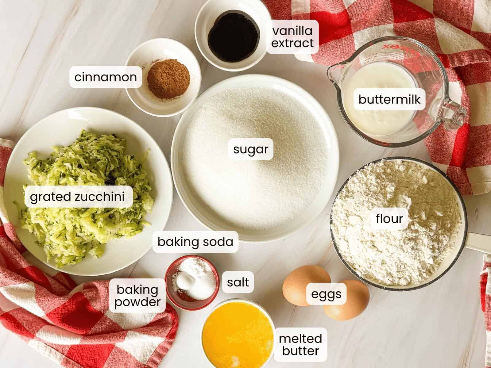 Baking ingredients for zucchini bread&mdash;including grated zucchini, sugar, flour, eggs, buttermilk, melted butter, vanilla extract, cinnamon, baking powder, baking soda, and salt&mdash;arranged on a countertop.