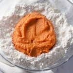 A glass bowl containing flour with a portion of orange sweet potato puree placed in the center, ready for mixing—perfect for creating tender pumpkin buttermilk biscuits.