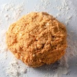A ball of orange-colored dough for pumpkin buttermilk biscuits rests on a floured marble surface.