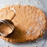 Biscuit dough for pumpkin buttermilk biscuits is rolled out on a floured surface, with a metal biscuit cutter pressing out a round shape.
