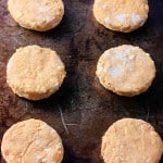Eight unbaked pumpkin buttermilk biscuits are arranged in rows on a dark, weathered baking sheet.