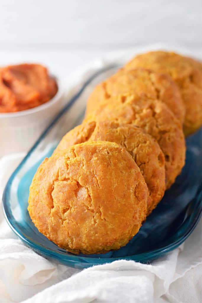 Five golden brown pumpkin buttermilk biscuits are arranged in a row on a blue glass plate, with a small bowl of reddish spread in the background.