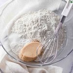 A glass bowl with flour, brown sugar, and salt next to a metal whisk, set on a white cloth—perfect for preparing pumpkin buttermilk biscuits.