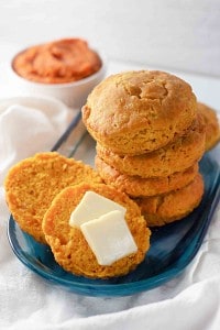 A stack of sweet potato biscuits and pumpkin buttermilk biscuits sits on a blue plate, with one biscuit sliced open and topped with two pats of butter; a bowl of orange spread is in the background.