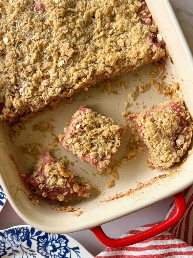 A baking dish with oat-topped dessert bars, three pieces cut and separated, showing a pink fruit filling, on a white kitchen surface.