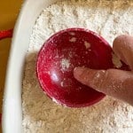 A hand presses a red measuring cup into a dish filled with flour and oats, flattening the mixture.