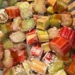 Chopped rhubarb pieces coated with granulated sugar in a mixing bowl, with a wooden spoon partially visible.
