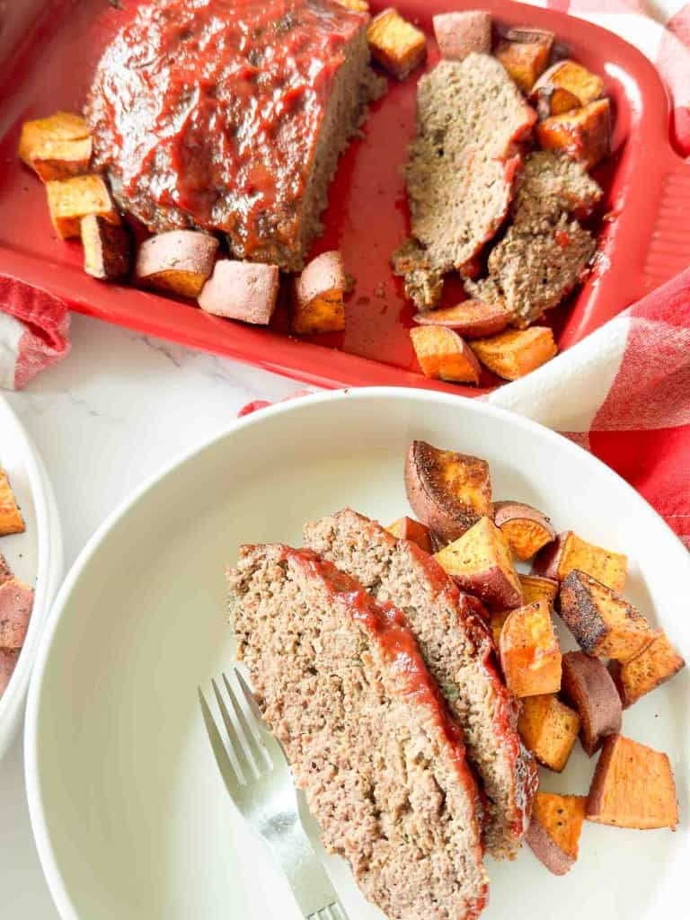 Sliced meatloaf with ketchup glaze served with roasted sweet potatoes on a white plate; more meatloaf and potatoes on a red baking dish in the background.