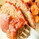 A close-up of a fork holding a piece of meatloaf with ketchup and a chunk of sweet potato, with slices of meatloaf and sweet potatoes in the background.