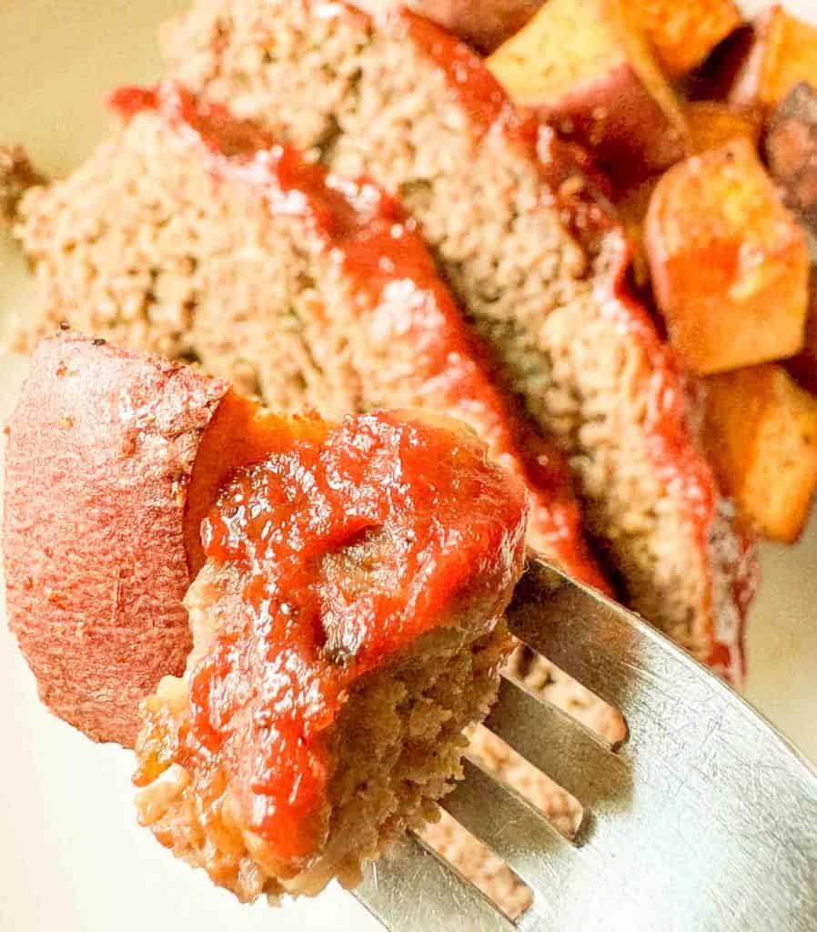 A close-up of a fork holding a piece of meatloaf with ketchup and a chunk of sweet potato, with slices of meatloaf and sweet potatoes in the background.
