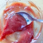 A metal spoon mixing red and beige sauce in a glass bowl.