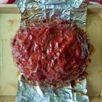 A cooked meatloaf covered in red sauce sits on a baking sheet lined with aluminum foil.