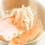 A close-up of a pink spatula holding a scoop of orange frosting, destined for a snickerdoodle cookie cake, with more frosting in a metal bowl in the background.
