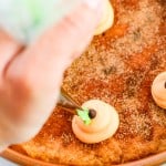 Hand piping green frosting onto a round snickerdoodle cookie cake, dusted with cinnamon-sugar, with a close-up focus on the decorating process.