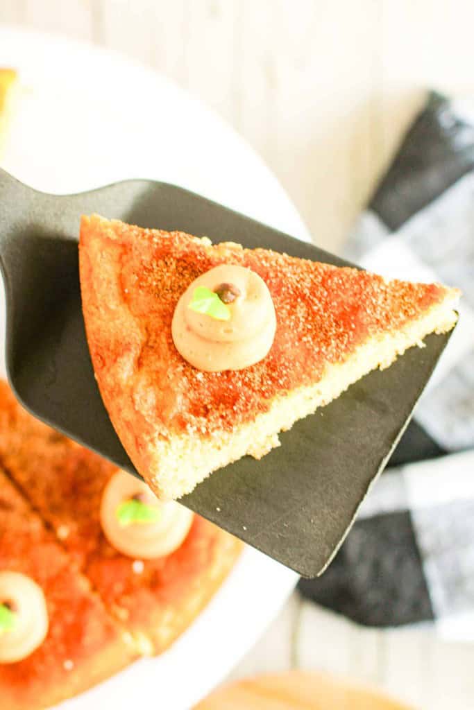 A slice of snickerdoodle cookie cake with frosting and a small green garnish is held on a spatula above a plate with more cake slices in the background.