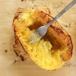 A fork scrapes cooked spaghetti squash inside its roasted shell for a delicious Spaghetti Squash Bake, with pepper and seasonings sprinkled on top, all on a parchment-lined baking sheet.