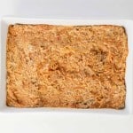 Rectangular white baking dish with a layer of mixed meat, sauce, and cheese—perfect for a comforting Spaghetti Squash Bake. Two small glass bowls with grated cheese are on either side of the dish on a white surface.