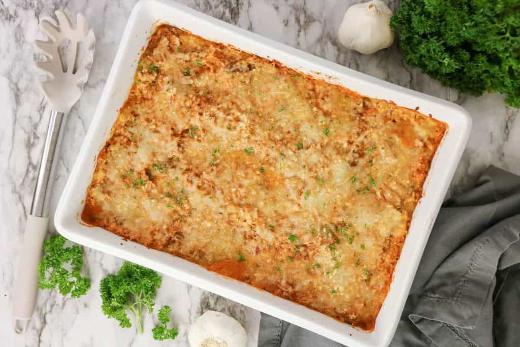 Baked lasagna in a white rectangular dish on a marble surface, garnished with chopped parsley, with kitchen utensils, garlic bulbs, and fresh parsley nearby—perfect for those craving classic comfort or considering a Spaghetti Squash Bake twist.