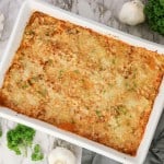 Baked lasagna in a white rectangular dish on a marble surface, garnished with chopped parsley, with kitchen utensils, garlic bulbs, and fresh parsley nearby—perfect for those craving classic comfort or considering a Spaghetti Squash Bake twist.