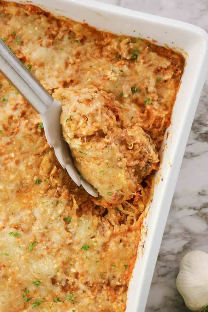 A Spaghetti Squash Bake casserole topped with melted cheese is being served from a white dish with tongs.