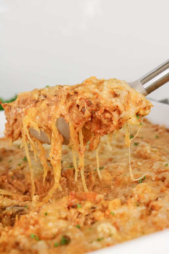 A serving spoon lifts a portion of cheesy, baked Spaghetti Squash Bake casserole from a dish, with melted cheese stretching from the pan.